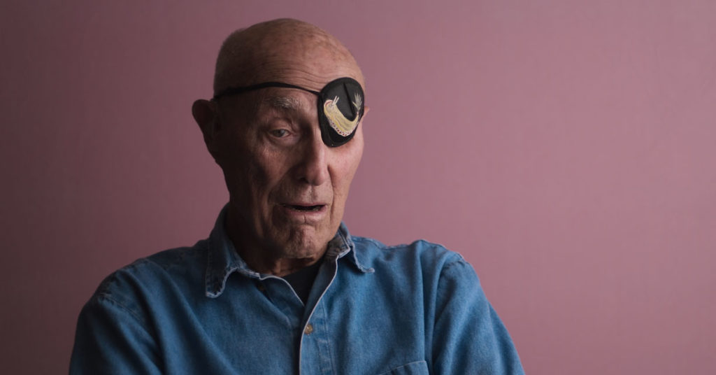 Brisbane-based colourist Angela Cerasi from Peachy Keen Colour graded this video content of an older, balding man wearing a blue demin shirt and an eyepatch, being interviewed against a pink backdrop, for the documentary "Playing With Sharks"