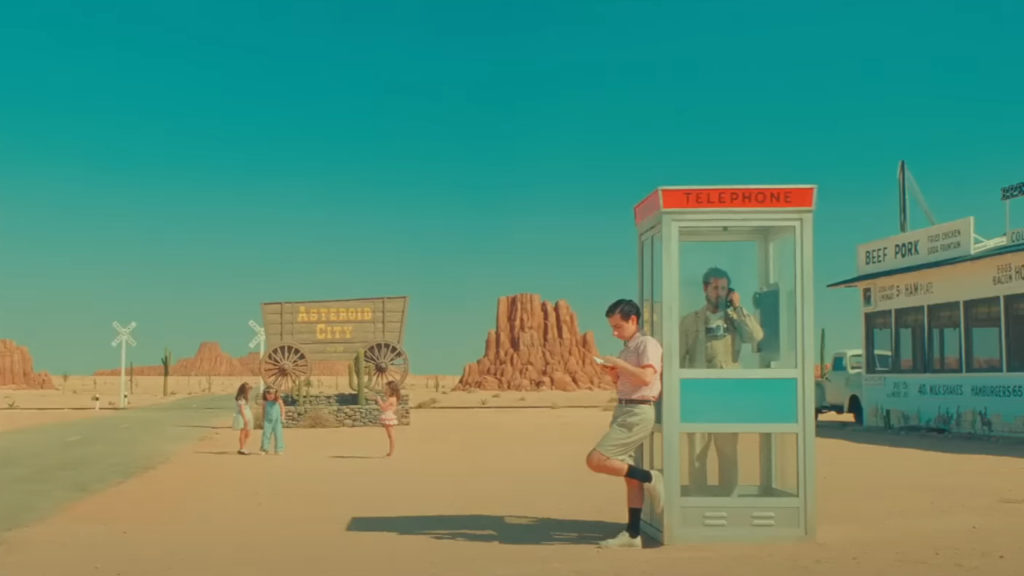 Green-tinged colour graded shot of a man leaning against a phone booth, people in a desert landscape in the background. Image from Wes Anderson's 2023 film "Asteroid City"