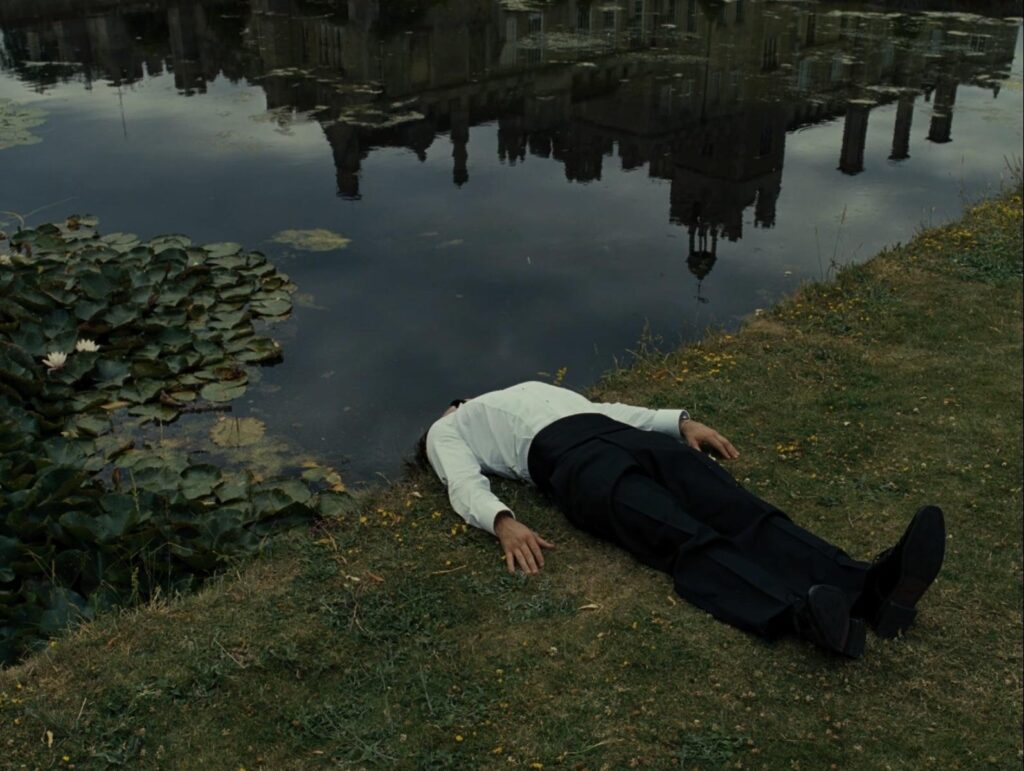 The visual language of Saltburn - image of a man lying on the grass, his head obscured by a slope leading into a pond. The image of a grand estate is reflected in the pond.